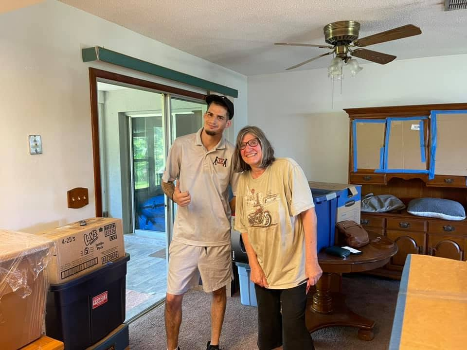 Two people smiling in a room during renovation with cardboard boxes and blue protective coverings on furniture